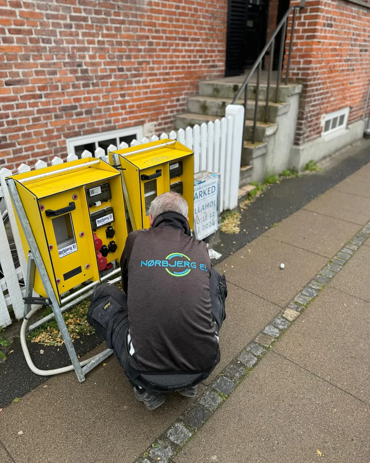Tekniker kobler to gule strømtavler ved et hvidt stakit på en gade